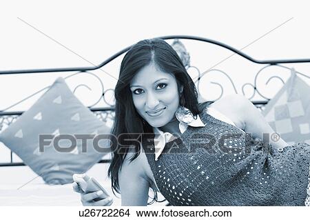 Picture - Portrait of a young woman lying on the bed and holding a mobile phone. Fotosearch