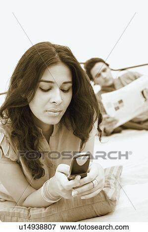 Young woman holding a mobile phone and a young man reading a newspaper in the background View Large Photo Image Stock Photo - Young woman holding a mobile phone and a young man reading a newspaper in the background. Fotosearch