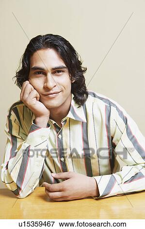 Stock Photo - Portrait of a young man sitting at the table with his hand on his chin. Fotosearch