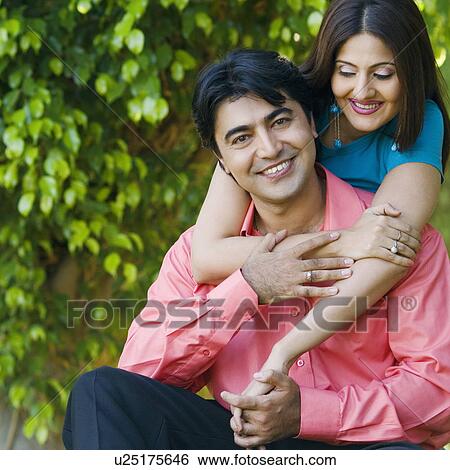 Stock Photograph - Close-up of a young woman embracing a young man from behind in a lawn. Fotosearch