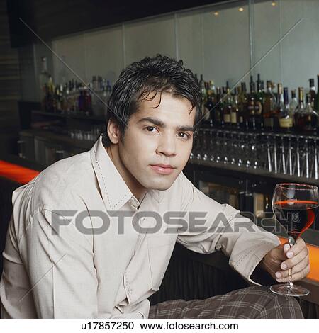 Stock Image - Portrait of a young man sitting at a bar counter and holding a glass of red wine. Fotosearch