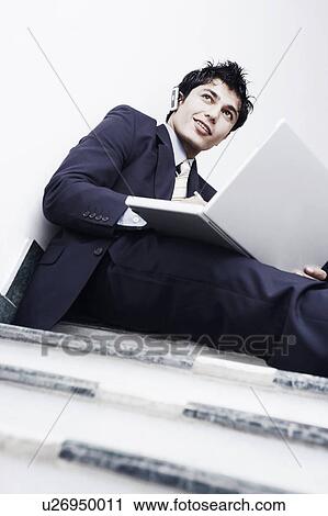 Stock Image - Low angle view of a businessman sitting on the staircase and using a laptop. Fotosearch