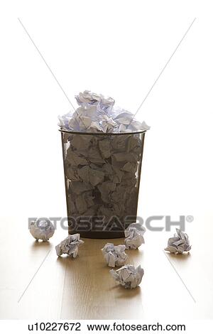 Stock Image - Full wire mesh trash can with crumpled paper scattered around. Fotosearch