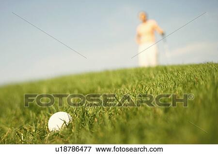 Stock Photo - Close-up of a golf ball in grass with a woman in the background. Fotosearch