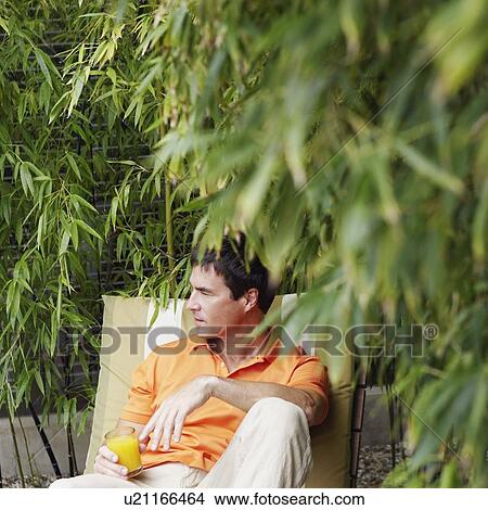 Picture - Close-up of a mature man reclining on a lounge chair and holding a glass of juice. Fotosearch