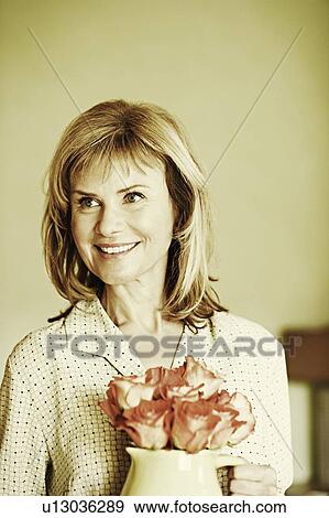 Stock Photo - Close-up of a mature woman holding a pitcher with flowers and smiling. Fotosearch