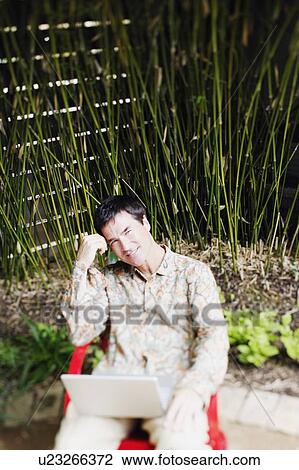 Stock Image - Portrait of a mature man sitting on a chair with a laptop on his lap. Fotosearch
