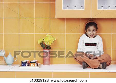 Stock Image - Portrait of a girl sitting on a kitchen counter. Fotosearch