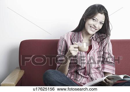 Stock Photograph - Young woman sitting on a couch holding a cup of tea and a magazine. Fotosearch