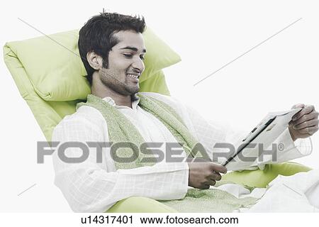 Close-up of a young man sitting on a chair reading a magazine View Large Photo Image Stock Image - Close-up of a young man sitting on a chair reading a magazine. Fotosearch