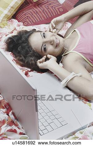 Stock Image - Portrait of a young woman lying on the bed with a laptop beside her. Fotosearch