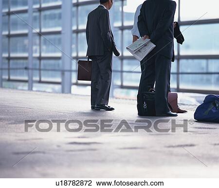 Stock Image - Three men in formal suit waiting on the platform with their bags and newspaper. Fotosearch