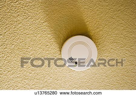 Stock Image - White smoke detector on yellow ceiling. Fotosearch