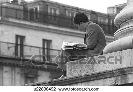 Stock Image - Man sitting at the base of a pillar reading a newspaper in Paris, France. Fotosearch
