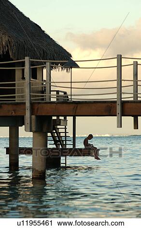 Stock Image - A man sitting on steps of a pier, Moorea, Tahiti, French Polynesia, South Pacific. Fotosearch