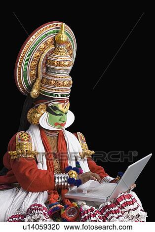 Stock Image - Close-up of a Kathakali dance performer sitting and using a laptop. Fotosearch