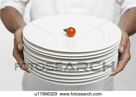 Stock Photo - Chef holding stack of dinner plates with single cherry tomato on top (mid section). Fotosearch