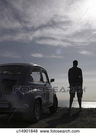 Stock Photo - Man standing beside parked car at beach, looking at ocean horizon, rear view. Fotosearch