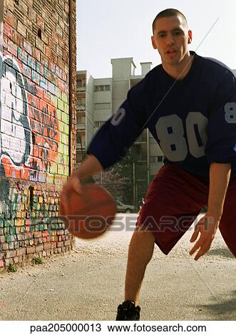 Stock Image - Man dribbling basketball in urban playground next to graffitied wall. Fotosearch