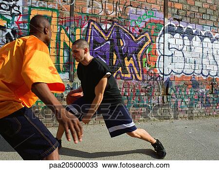 Stock Image - Man dribbling basketball, opponent blocking, next to graffitied wall. Fotosearch