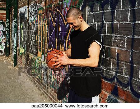 Picture - Man leaning on graffitied wall, looking down, holding basketball. Fotosearch