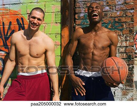 Stock Image - Two shirtless men standing and laughing next to graffitied wall, one holding basketball. Fotosearch