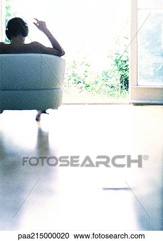 Stock Image - Man sitting on sofa, listening to headphones, rear view.. Fotosearch