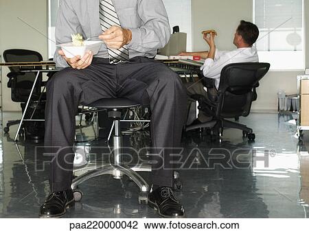 Stock Image - Two businessmen in office, eating.. Fotosearch