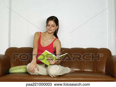 Stock Image - Woman sitting on sofa, holding book. Fotosearch