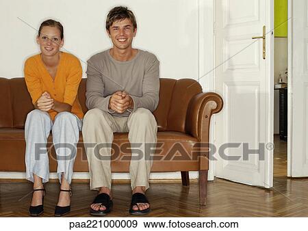 Stock Photo - Young man and woman sitting side by side on sofa, off center. Fotosearch