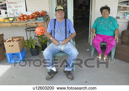 Stock Image - Man and wife sits in chair in front of small vegetable and produce store. Fotosearch