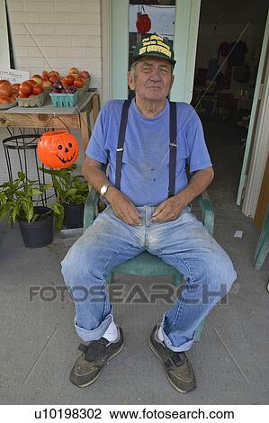 Stock Image - Man sits in chair in front of small vegetable and produce store. Fotosearch