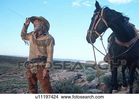 Picture - Senior cowboy standing with horse in desert. Fotosearch
