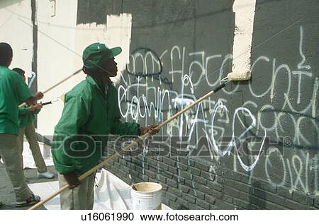 Stock Image - Three black youths painting over graffiti covered wall. Fotosearch