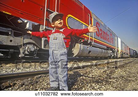 Stock Image - Little boy in engineers overalls and cap in front of train station. Fotosearch