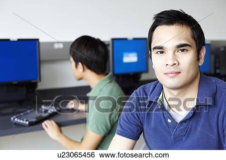 Portrait of a young man smirking with a young man sitting behind him and using a computer View Large Photo Image Stock Photograph - Portrait of a young man smirking with a young man sitting behind him and using a computer. Fotosearch