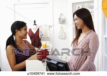 Stock Image - Side profile of two young women standing at a checkout counter and looking at each other. Fotosearch