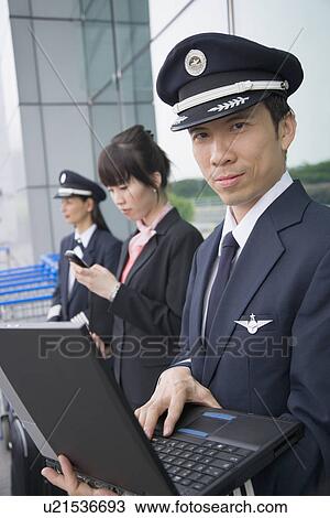 Stock Image - Side profile of a female cabin crew holding a mobile phone and standing with two pilots at an airport. Fotosearch