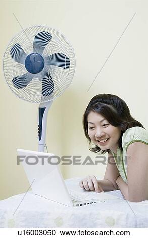 Stock Image - Side profile of a young woman lying on the bed and using a laptop. Fotosearch