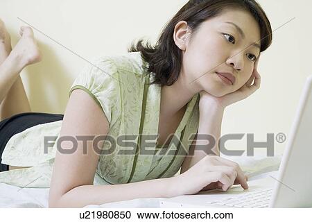 Stock Image - Side profile of a young woman lying on the bed and using a laptop. Fotosearch