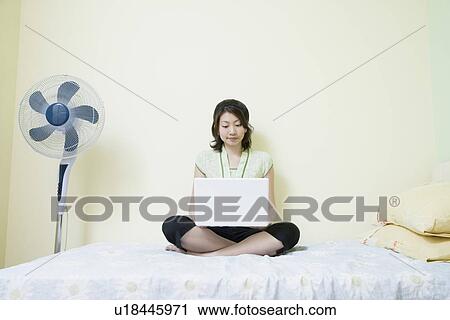 Stock Image - Young woman sitting cross-legged on the bed and using a laptop. Fotosearch
