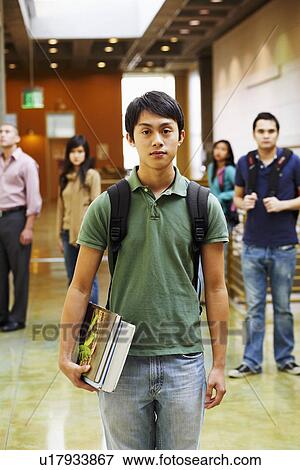 Stock Photo - Portrait of a young man holding books with four people standing behind him in the corridor. Fotosearch
