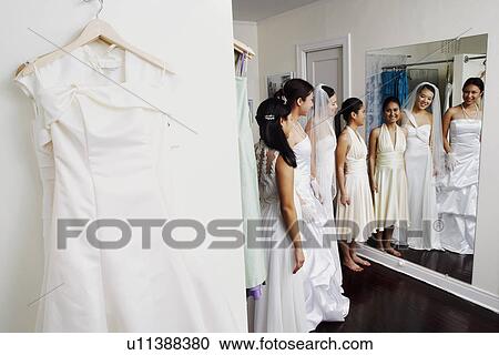 Three young women wearing wedding dresses and looking at themselves in a mirror View Large Photo Image Stock Image - Three young women wearing wedding dresses and looking at themselves in a mirror. Fotosearch