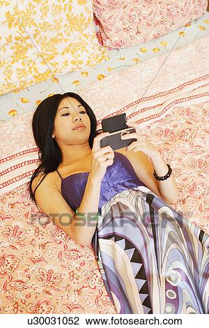 Stock Image - High angle view of a young woman lying on the bed and using a mobile phone. Fotosearch
