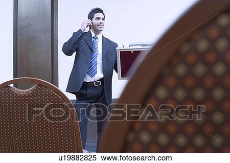 Businessman standing in a conference room and talking on a mobile phone View Large Photo Image Stock Photography - Businessman standing in a conference room and talking on a mobile phone. Fotosearch