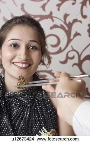 Picture - Close-up of a person's hand feeding a young woman with chopsticks. Fotosearch