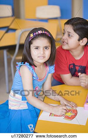 Stock Photo - Girl drawing on a sheet of paper with a boy sitting beside her in a classroom. Fotosearch