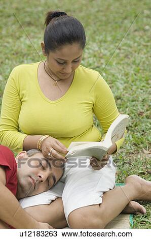 Stock Image - High angle view of a mid adult woman reading a book with a mid adult man lying on her lap . Fotosearch