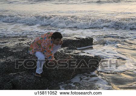 Stock Image - High angle view of a young man crouching on rocks on the beach. Fotosearch