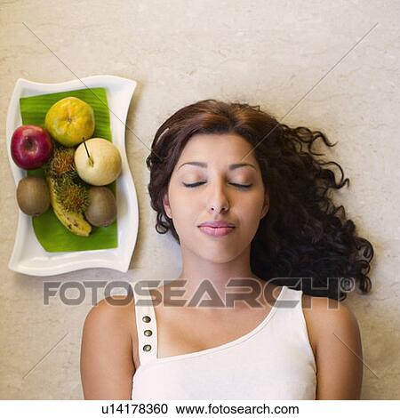 Stock Image - High angle view of a young woman lying near a fruit tray. Fotosearch